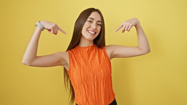 Cheerful young woman in sleeveless t-shirt standing proud in a yellow room, confidently pointing at herself, looking happy and successful