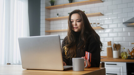 Asian woman enter credit card number on laptop for shopping online, makes secure easy distant electronic payment while sitting in kitchen at home	