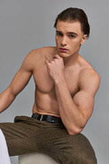 enticing shirtless man in brown pants sitting and looking alluringly at camera on gray backdrop