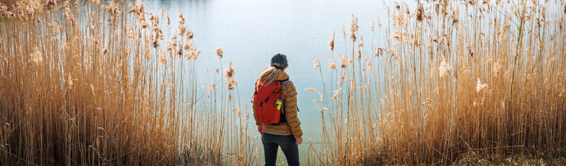 Woman with red backpack hiking at lake with reed grass. Symmetrical panoramic view of nature and a hiker enjoying time outdoors