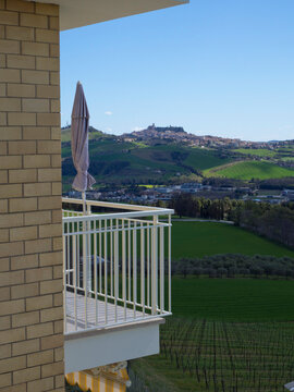 Scenic view from a balcony with a closed sun umbrella, of green hills, vineyards and the town of Fermo in the background; Monte Urano, Marche, Italy