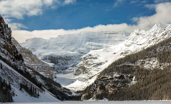 Scenic view of snow covered mountains and glacier with blue sky and clouds overshadowing the snow covered lake; Lake Louise, Alberta, Canada