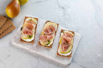 Ideal snack wholewheat crispbread with ricotta cheese, prosciutto, sliced pear and thyme on light grey marble cutting board. Healthy snack concept. Italian cuisine