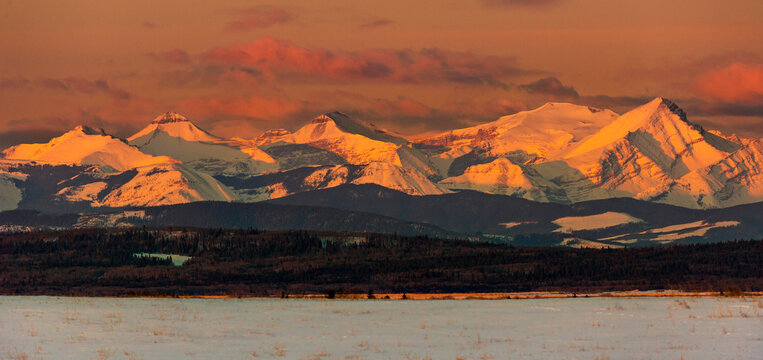 Snow-covered mountain range glowing with the warm light of sunrise and warm glowing clouds with foothills and snow-covered field in the foreground, West of Calgary, Alberta; Alberta, Canada