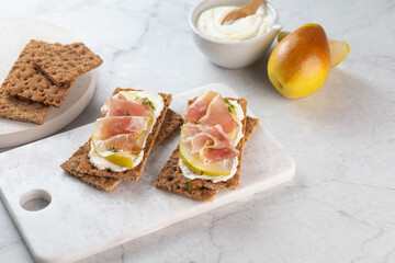 Ideal snack wholewheat crispbread with ricotta cheese, prosciutto, sliced pear and thyme on light grey marble cutting board. Healthy snack concept. Italian cuisine