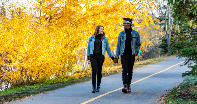A mixed race married couple smiling at each other, walking down a road in a city park during a fall family outing, spending quality time together; Edmonton, Alberta, Canada