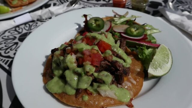 carne asada vampiro tostada and fish taco on a plate at a mexican restaurant (green avocado sauce, lime, white plate) garnished with radishes, jalapeno peppers, pico de gallo, tomatos onions cilantro
