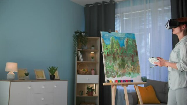 augmented reality, modern creative female artist uses viar glasses to draw a picture on canvas with paints and brushes while standing in a cozy room
