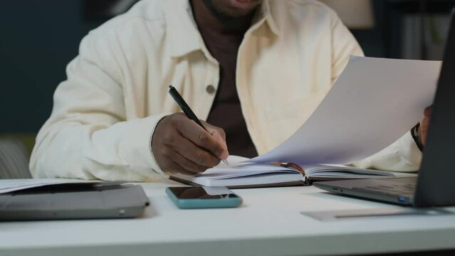 Cropped shot of unrecognizable Black businessman examining financial report and taking notes in his notebook while working at office desk at night