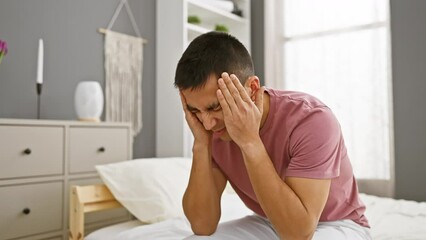 A pained young hispanic man sitting on a bed indoors holding his head as if experiencing a headache or stress.