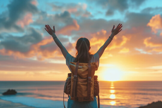 Close up back view of young woman with a travel backpack on her back and holds her hands up as a sign of freedom standing ar seaside at sunset. Joyful free travel concept