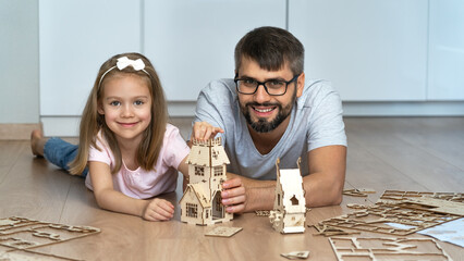 Happy father and child look at camera and laugh building wooden construction set. Family play with...