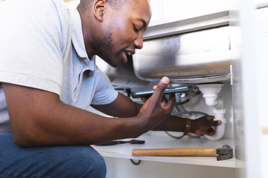 African American plumber fixing sink while making phone call