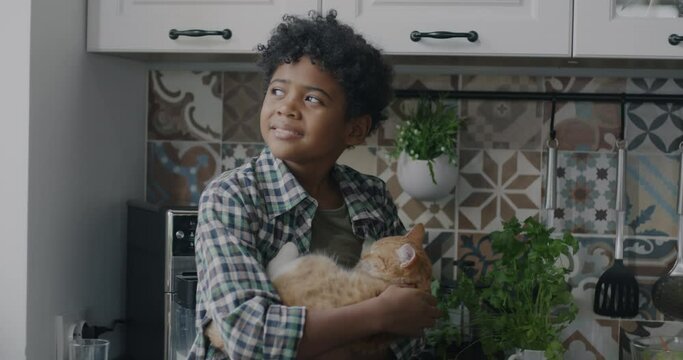 Slow motion portrait of adorable African American kid sitting on kitchen table and holding kitten stroking animal. Childhood and pets concept.