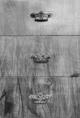 The front of an old wooden chest of drawers with scratches and imperfections with three carved metal handles. Black and white photo