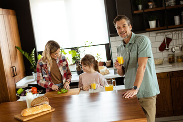 Joyful man with a glass of orange juice while a woman and child prepare food in a cozy kitchen - 763446519