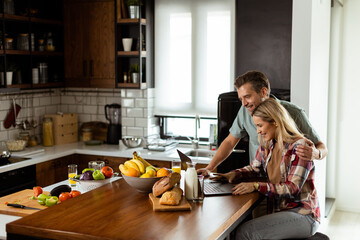 Cheerful couple enjoys a light-hearted moment in their sunny kitchen, working on laptop surrounded by a healthy breakfast - 763446787