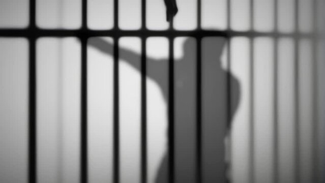 The silhouette of a lone boxer, wearing a hoodie, training in solitude, projects its shadow onto a white wall, with the overlay of prison bar shapes.
