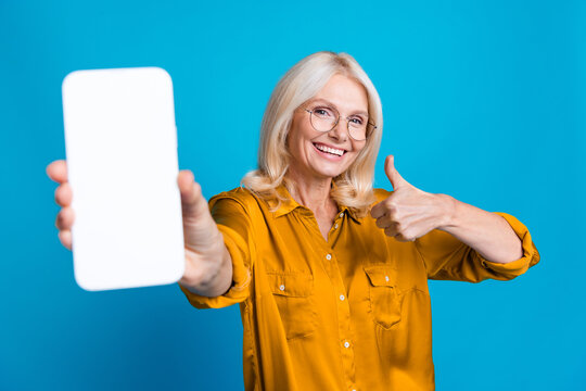 Photo portrait of lovely senior lady hold gadget white screen thumb up wear trendy yellow silky garment isolated on blue color background