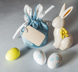 Easter image. Creative gift bag with bunny ears, blank note, ceramic bunny, decorative eggs on gray.