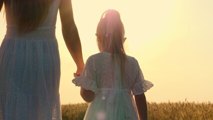 Mother and daughter walking at sunny autumn wheat field holding hands with love tenderness back...