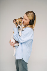 Blonde woman with big black headphones and blue shirt holding cute dog Jack Russell terrier. Studio shot. Grey background. Listening to music with pet