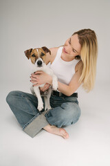Cute blonde woman looking at the dog sitting on her lap. Studio portrait. Dog owner love. Looking at camera. Grey background. Blue jeans white shirt