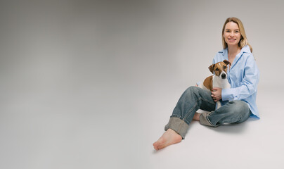 Smiling blonde woman in a blue shirt sits on the floor, the dog sitting on her laps. Funny cute friends studio portrait. Grey background. Large size banner