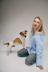 laughing blonde woman in a blue shirt sits on the floor, the dog stands with its paws on her leg. Funny cute friends studio portrait. Grey background. 