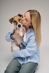 Kissing a pet. Blonde beautiful woman in blue shirt holding her dog Jack Russell terrier and kissing. Studio shot. Grey background