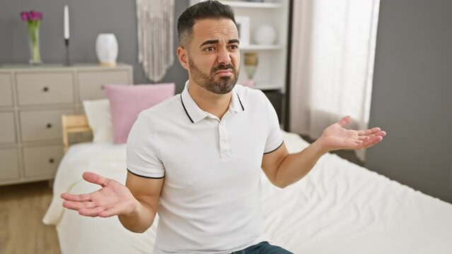 Clueless young hispanic man sitting on bed, with puzzled doubting expression in his bedroom