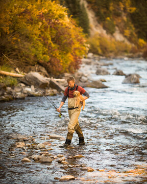 Man carries his puppy across the river while fishing in the fall on the river