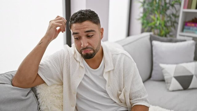 Pensive young arab man, sitting alone on home sofa, lost in thought, confused and unsure, wrestling with a mind-boggling question. a tableau of doubt, indoor contemplation concept.