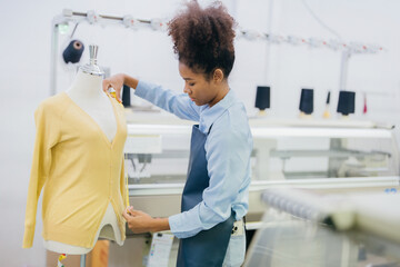 American female designer is using a tape measure on a mannequin to design a garment in a sewing industry. Around there are machines working and spools of thread for making shirt material.