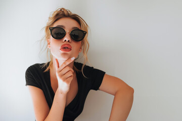 jeune femme mannequin envoyant des baisers, portant des lunettes de soleil noires, la main sur le menton, posant pour la photo, sur fond blanc dans le style de la photographie de mode - obrazy, fototapety, plakaty