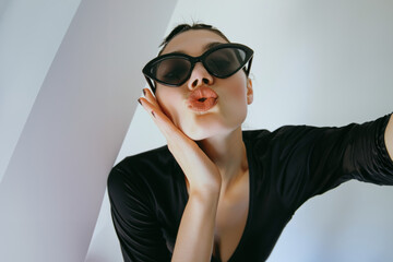 jeune femme mannequin envoyant des baisers, portant des lunettes de soleil noires, la main sur le menton, posant pour la photo, sur fond blanc dans le style de la photographie de mode
