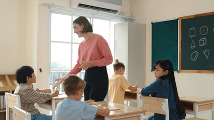 Caucasian teacher walking and check student homework at classroom while diverse children doing...