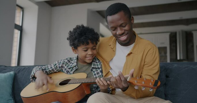 Loving father African American man teaching kid to play the guitar having fun in apartment together. Family and creative activity concept.