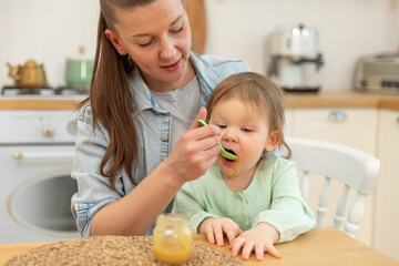 Obraz na płótnie Canvas Happy family at home. Mother feeding her baby girl from spoon in kitchen. Little toddler child with messy funny face eats healthy food at home. Young woman mom giving food to kid daughter