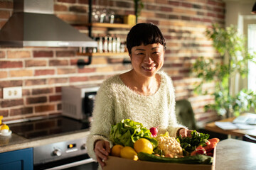 Woman taking vegetables out of cardboard box in kitchen at home - obrazy, fototapety, plakaty