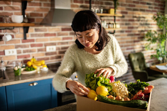 Woman taking vegetables out of cardboard box in kitchen at home