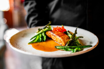 A close-up of a person holding a plate with grilled salmon, asparagus, and orange sauce. The dish is beautifully presented, with glossy salmon and grill marks on the asparagus