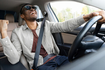 Joyful African American man in green glasses dances in the car, sings while driving. Road fun