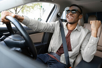 A joyful african american in glasses dances in a car, sings while driving his car. Road fun - obrazy, fototapety, plakaty