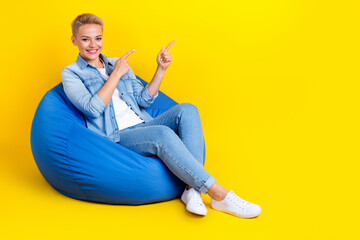 Full body photo of attractive young woman point empty space sit bean bag wear trendy jeans clothes...