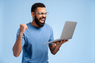 Handsome African American excited man wearing eyeglasses, holding computer, using laptop, win money
