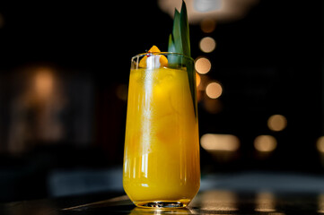 refreshing yellow cocktail with a green leaf garnish, served in a tall glass against a dark, bokeh background. The drink appears cold and enticing, and the lighting highlights its vibrant color.