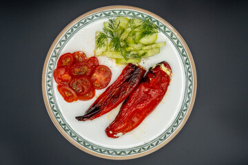 Grilled pointed peppers, filled with spicy feta cheese, garnished with lettuce and cucumber slices with dill and cocktail tomatoes on a plate. Shot from above with a dark background