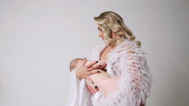 Beautiful woman with long blond hair wearing white robe waving a baby in hands. Mother and child concept. White backdrop.