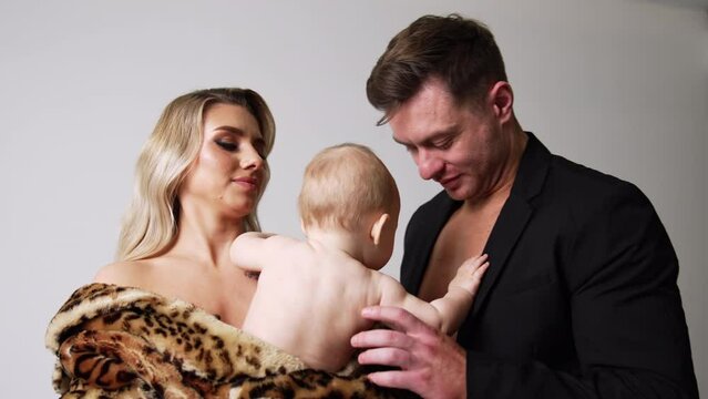 Blonde woman wearing fur coat holding a little naked baby. Man in black jacket kisses and embraces the child.